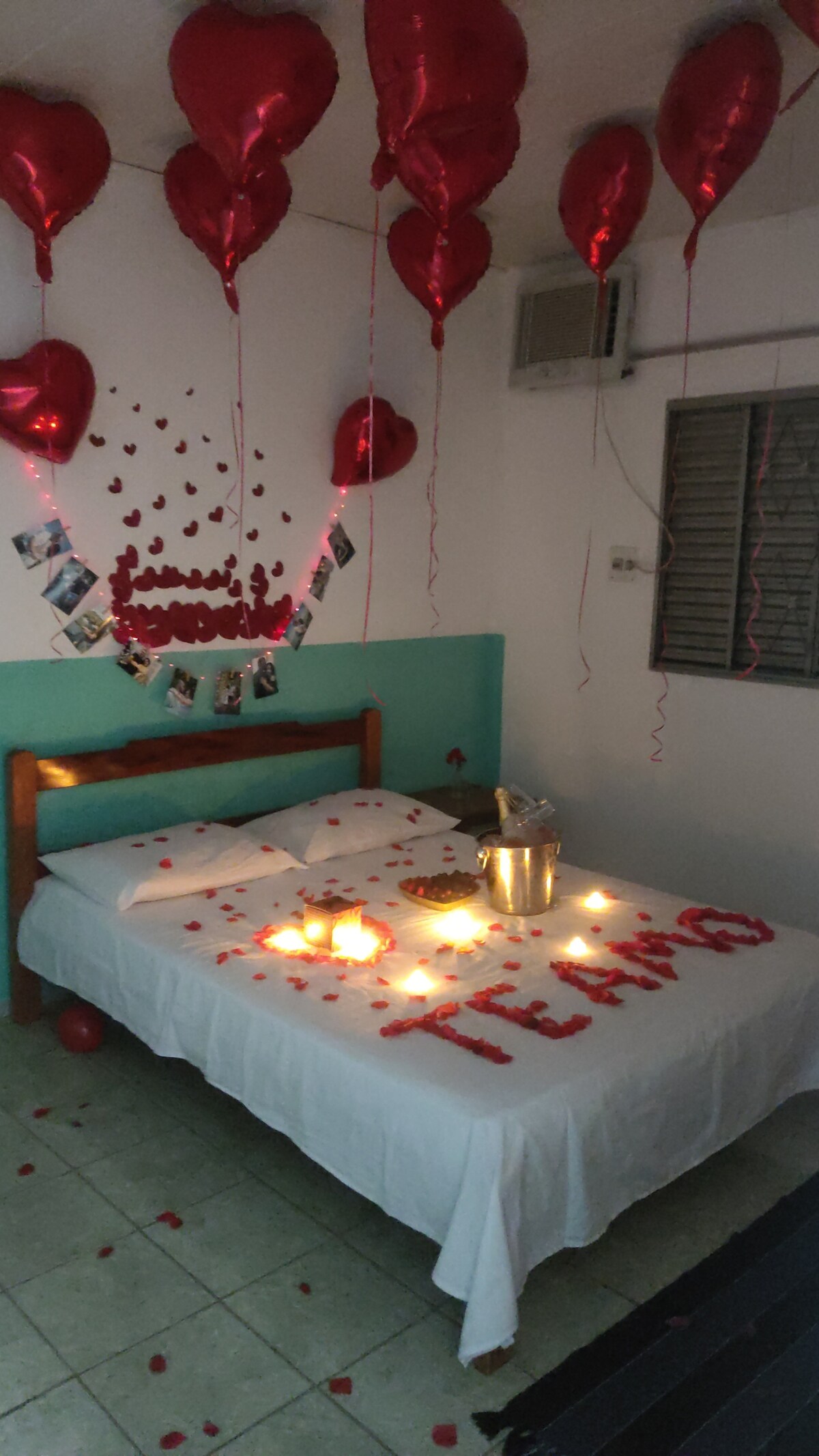A romantic bedroom is adorned with red balloons and heart decorations above a spacious bed. Red rose petals are scattered across the white bedding, and candles are arranged nearby, creating a cozy ambiance for a special occasion.