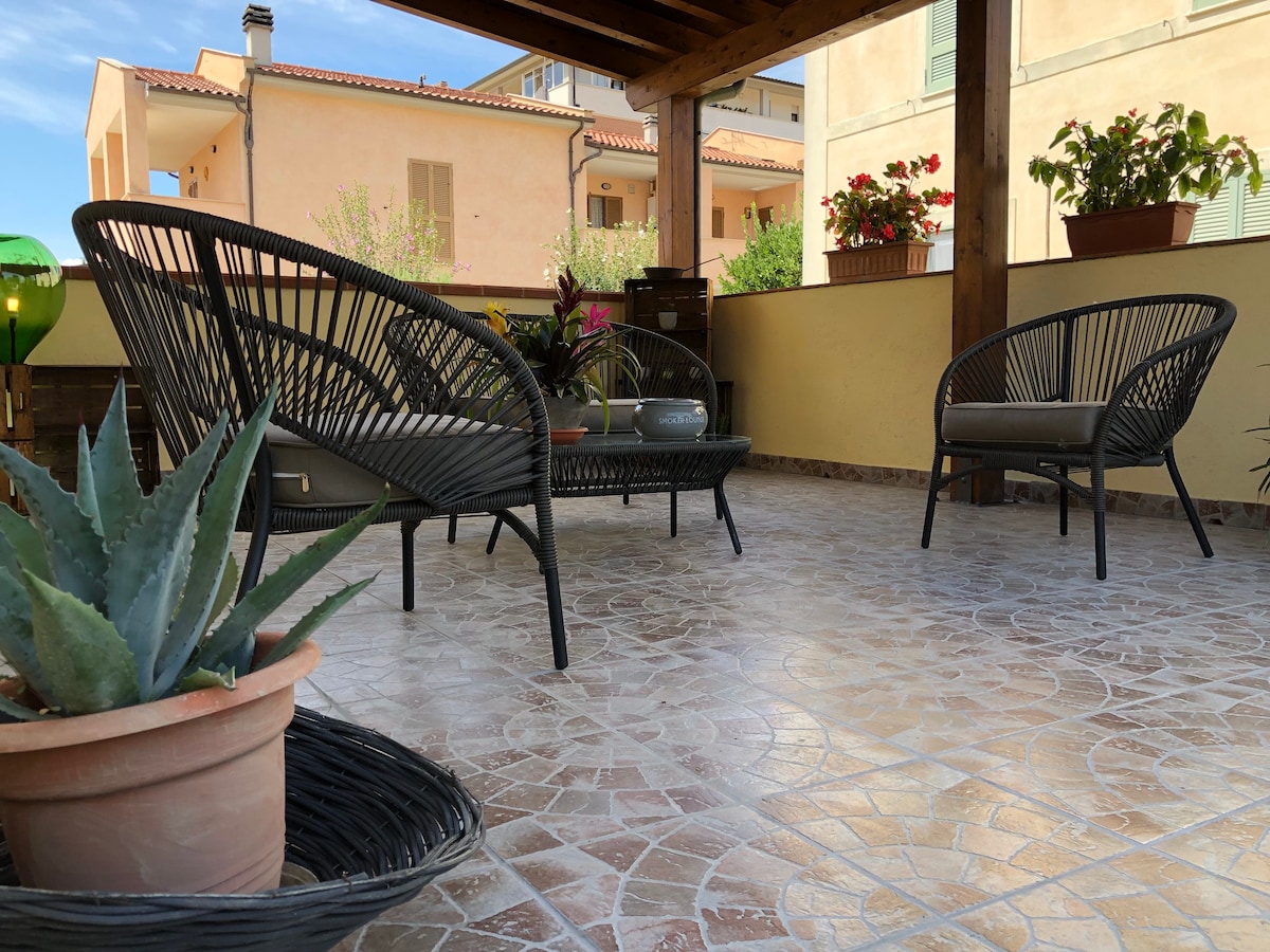 A spacious terrace is showcased, featuring two black woven chairs and various potted plants. The tiled floor extends throughout the area, and light filters in from the surrounding buildings, contributing to a bright and inviting space for relaxation.