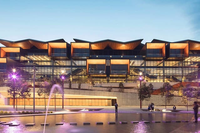 A modern building showcases a striking architectural design with a distinctive roofline. The exterior features large glass windows and warm wooden accents. Surrounding greenery and a water feature are visible in the foreground, contributing to a vibrant and inviting atmosphere.