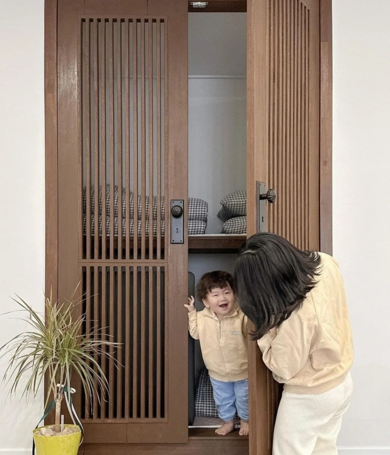 A unique storage space featuring wooden slatted doors opens to reveal neatly organized bedding. A child stands inside, while an adult leans down to interact. A potted plant adds a touch of greenery to the scene, enhancing the inviting ambiance of the area.