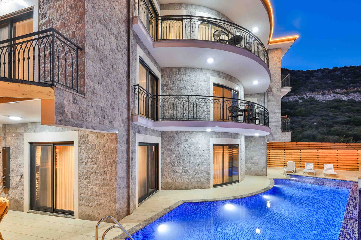 A modern building showcases multiple balconies overlooking a clear blue pool. The exterior features textured stone walls, and warm lighting highlights the architectural details. Lounge chairs are positioned by the pool, creating a relaxing ambiance in a serene environment.