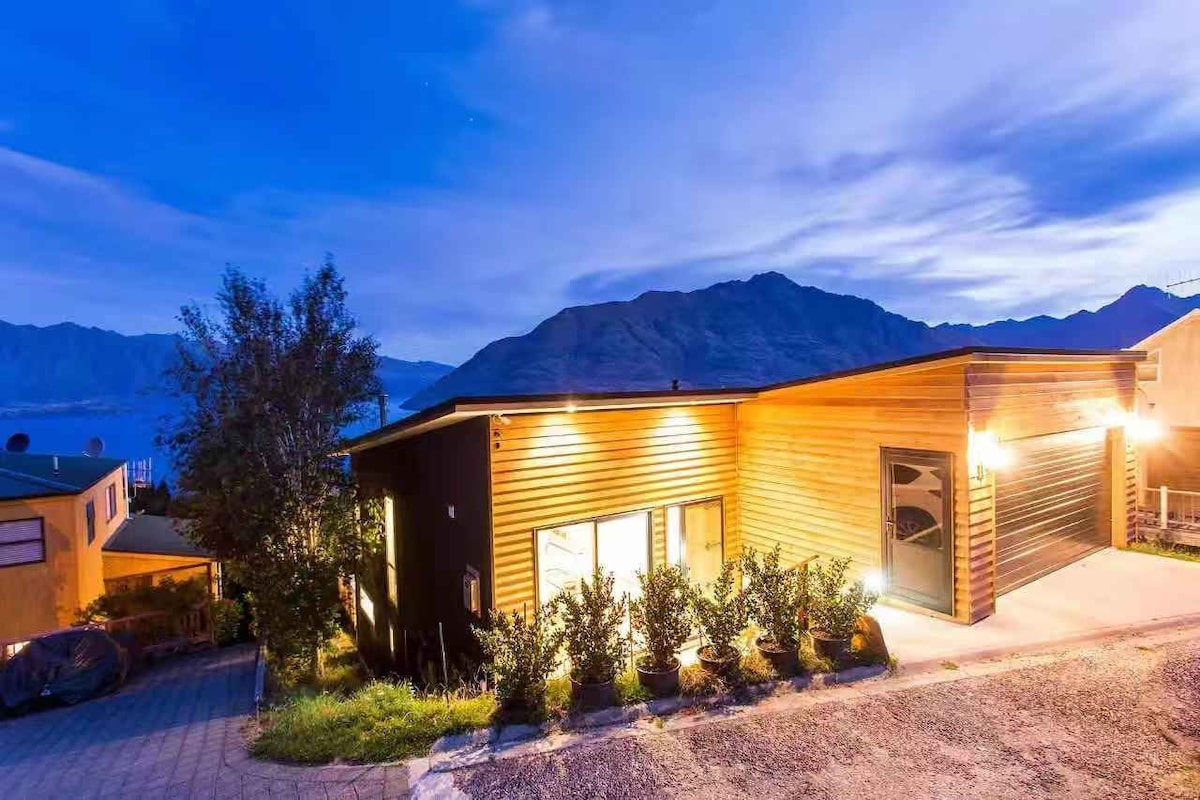 A modern holiday house is visible at dusk, featuring a wooden exterior illuminated by exterior lighting. The backdrop includes distant mountains, and the layout presents a welcoming entrance with landscaped shrubs lining the pathway.