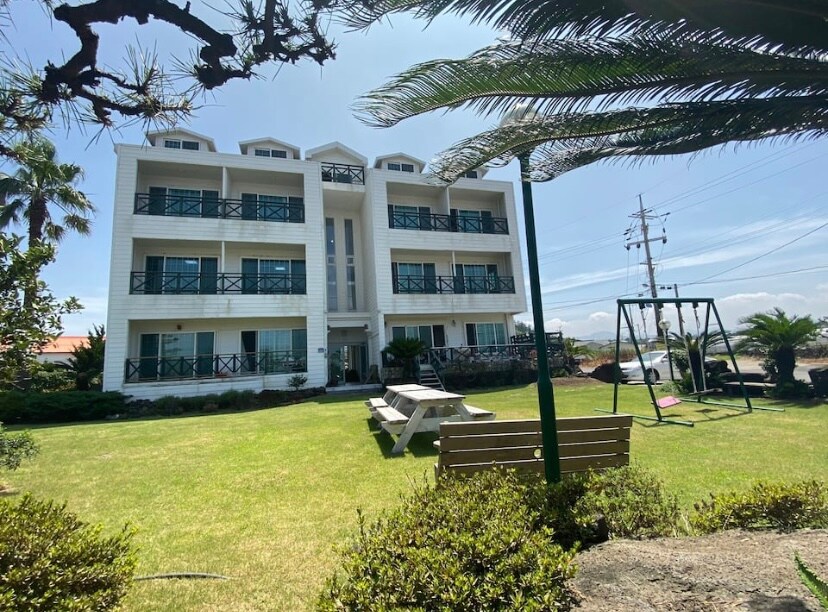 A three-story building with a modern design, featuring large balconies and multiple windows. The surrounding area is adorned with greenery and offers a grassy lawn, a picnic table, and a swing set, inviting outdoor activities and relaxation.
