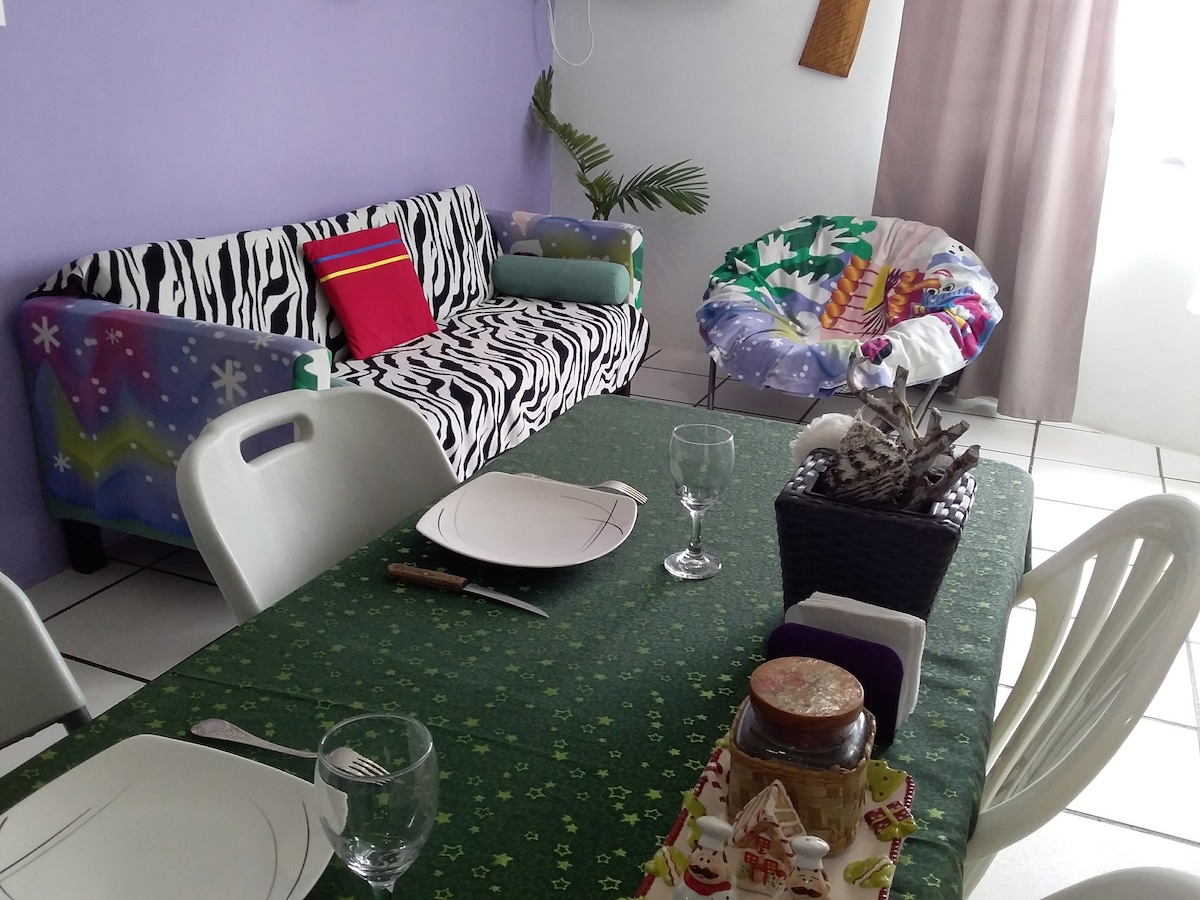 A bright dining area features a green tablecloth covering a rectangular table set for a meal, complete with plates and glasses. A vibrant zebra-print sofa is positioned nearby, accompanied by a colorful chair. Natural light fills the space through partially open curtains.
