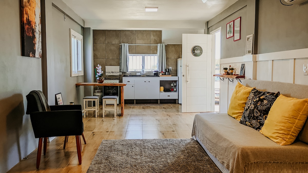 An open layout features a cozy seating area with a sofa and armchair, complemented by yellow and patterned cushions. The kitchenette is visible, showcasing modern appliances and ample counter space. Natural light streams through a window, enhancing the inviting atmosphere.
