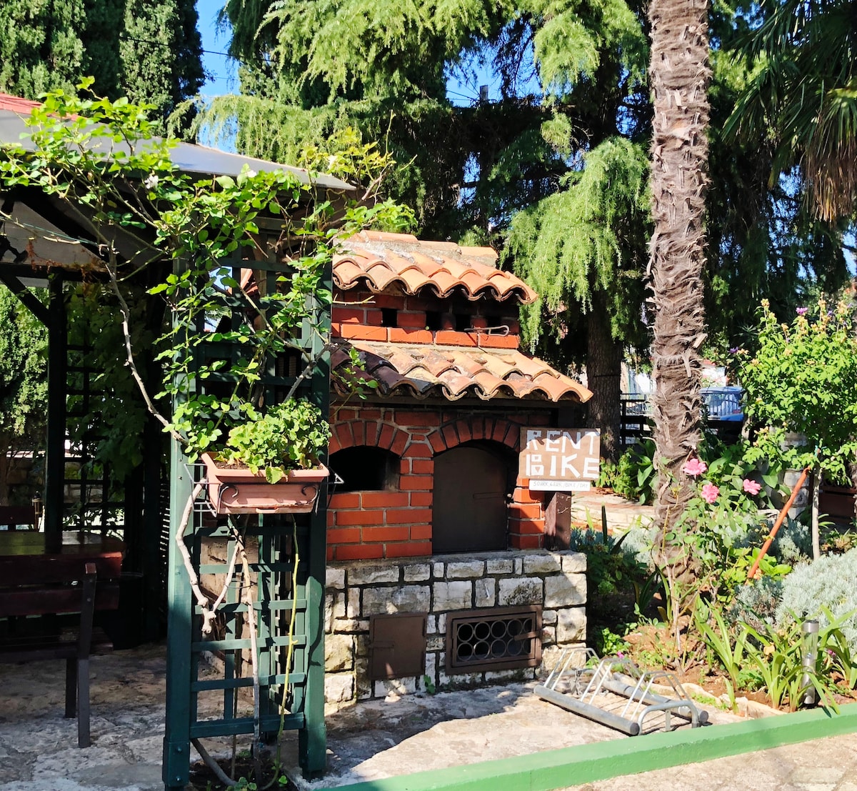 An outdoor area features a traditional brick barbecue oven surrounded by lush greenery. A gazebo is partially visible, providing a shaded spot. The landscaping includes palm and cypress trees, creating a natural and inviting environment for relaxation and outdoor cooking.
