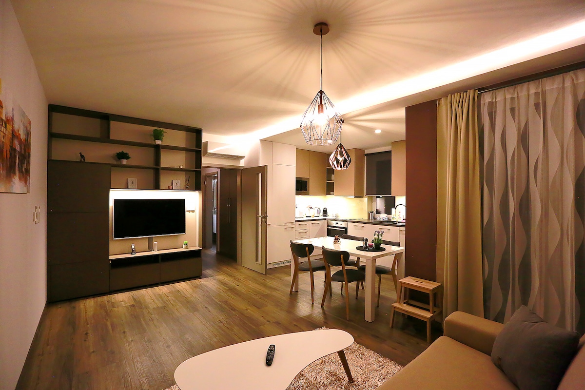 A modern living space features a spacious layout, highlighted by a dining area with a round table and four chairs. The open kitchen is showcased with sleek cabinetry and appliances. Soft lighting illuminates the room, creating a cozy atmosphere. A comfortable sofa is present in the foreground.