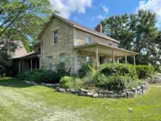The exterior of a spacious stone house is showcased, featuring a welcoming porch with a lush garden in front. Surrounding greenery and trees add to the serene atmosphere. The house exhibits a charming mix of historic stonework and modern elements under a clear blue sky.