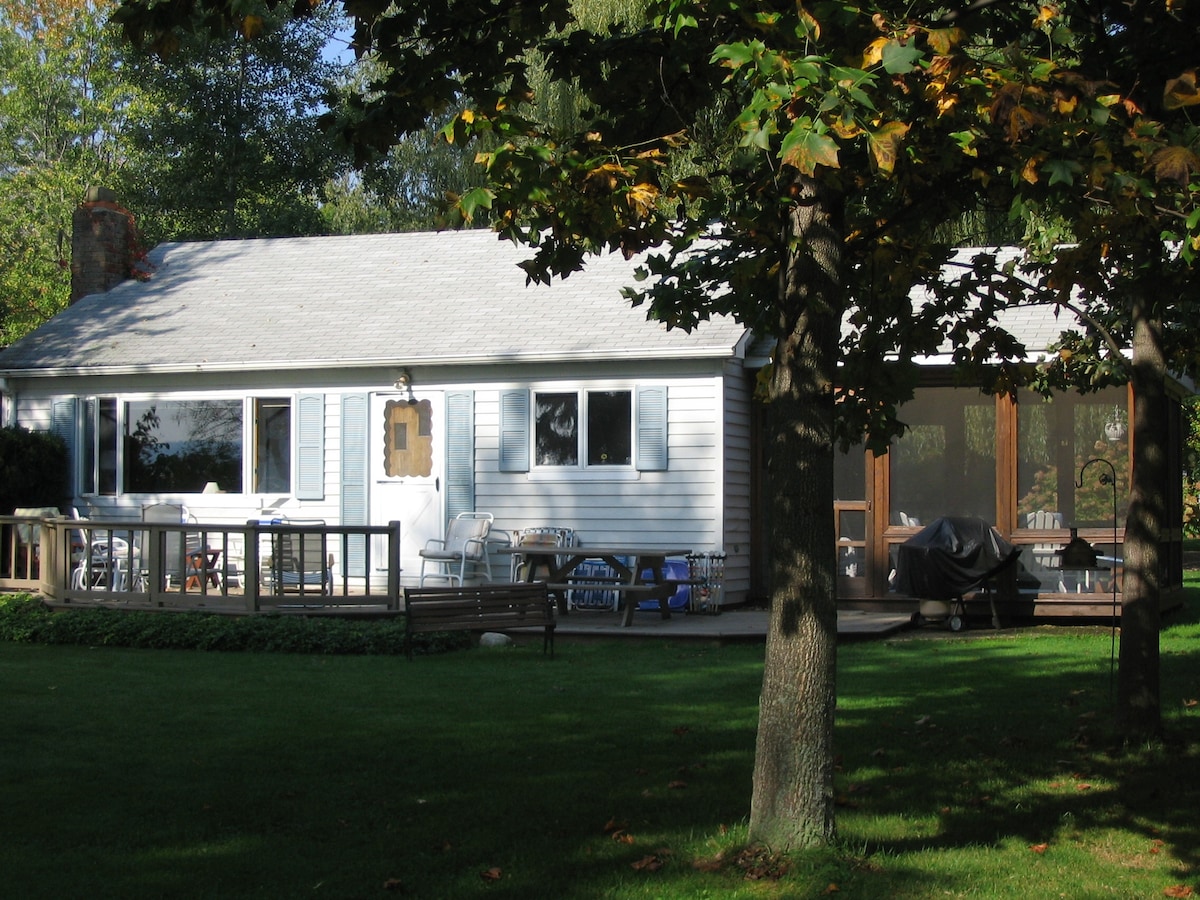 The cottage features a light-colored exterior with blue shutters and a spacious deck area. A large screened porch is visible on the right, providing a shaded space for relaxation. Green grass surrounds the cottage, and trees offer additional natural coverage.