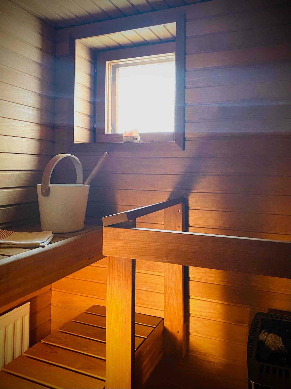 A wooden sauna interior is illuminated by natural light from a small window. A bench made of slatted wood is positioned beside a bucket, contributing to a soothing and functional environment. The textured wooden walls enhance the warm, inviting atmosphere.