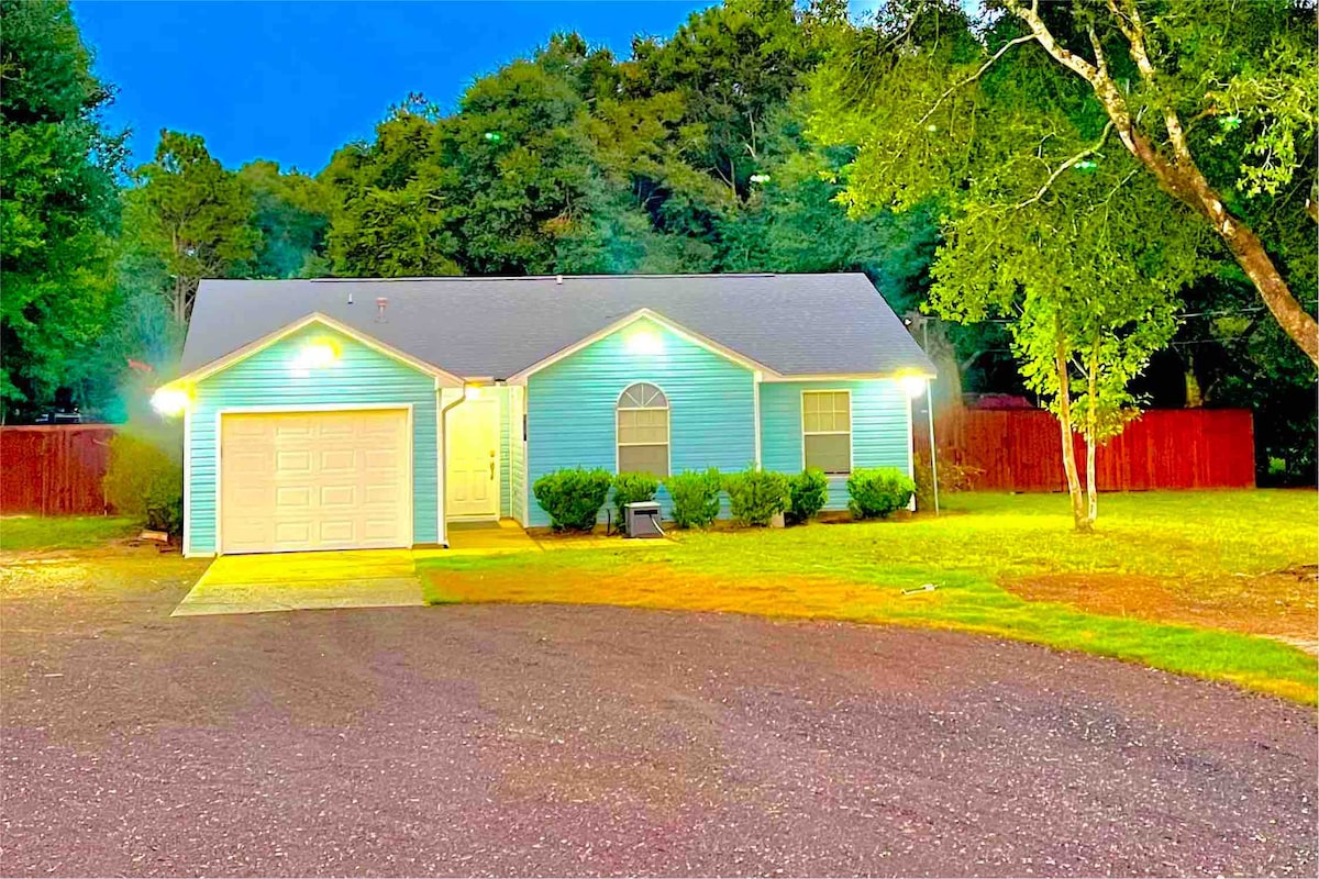 A single-story home is shown surrounded by trees, illuminated by exterior lights. A paved driveway leads to a garage, while lush green bushes frame the front. The welcoming blue exterior contrasts with the dark foliage in the background.