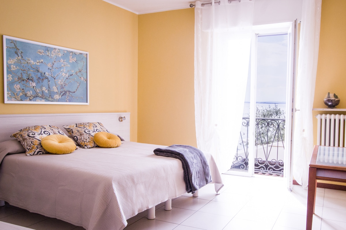 A bright bedroom features a spacious bed with decorative pillows. Clear curtains frame a door that leads to a small balcony, revealing a view of the lake. A simple wooden table is positioned beside the bed, complementing the warm yellow walls.