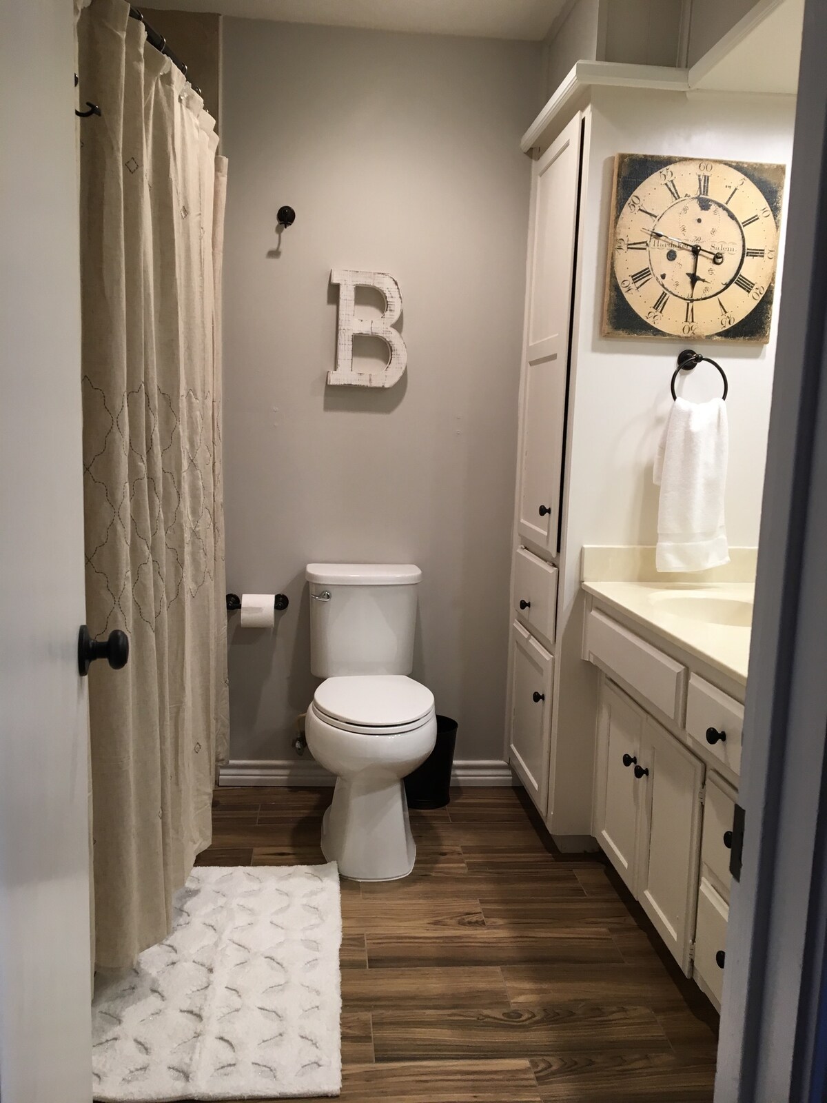 A modern bathroom is shown, featuring a beige shower curtain and a white tub. A toilet is positioned next to a wooden vanity with a light-colored countertop. Wall-mounted storage contains towels, and a clock adds a decorative touch. A textured rug is placed on the floor.