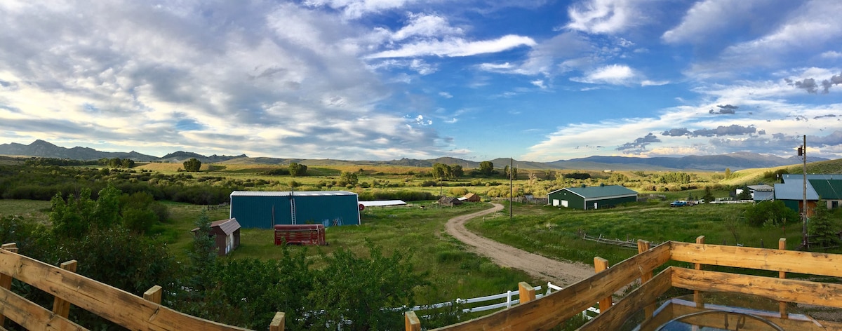 A panoramic view of the ranch landscape reveals expansive greenery and hilly terrain under a soft blue sky with scattered clouds. Dirt pathways meander through the property, while rustic buildings are visible among the natural surroundings, emphasizing the rural charm of the area.