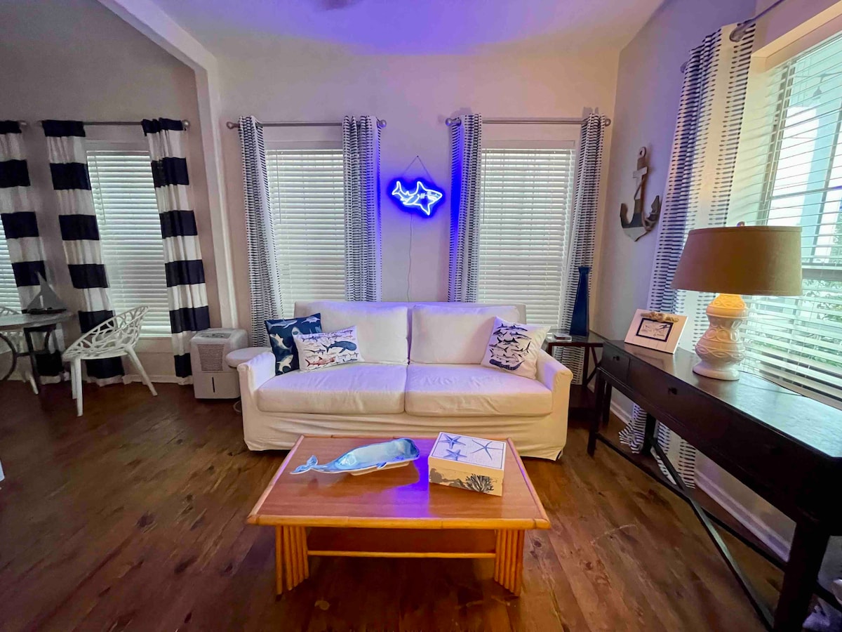 A light-filled living area features a white sofa adorned with blue and white pillows, complemented by wooden flooring. A wooden coffee table with nautical decor sits in front of the sofa. Large windows with sheer curtains allow natural light to brighten the space, enhancing the cozy atmosphere.