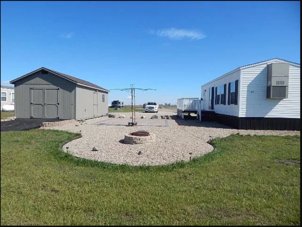 A spacious outdoor area is shown, featuring a central fire pit surrounded by gravel and grass. On one side, a gray shed stands, while a white trailer is visible on the opposite side. Open space surrounds the property, with vehicles parked in the background.