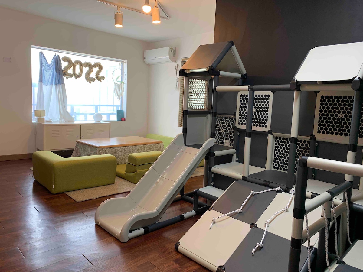 An engaging play area is designed for children, featuring a slide and climbing structure. A cozy seating arrangement includes green sofas and a coffee table, while playful decorations adorn the wall. Large windows allow natural light to brighten the space.