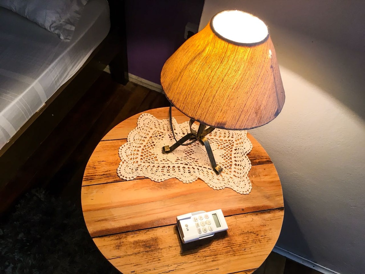 A rustic wooden bedside table is adorned with a delicate lace doily and a warm-toned lamp. A remote control is positioned beside the lamp. The background hints at a cozy sleeping area with a partially visible bed.