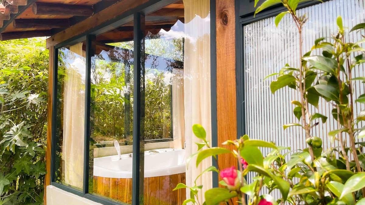 A private hot tub is visible through large glass windows, framed by natural greenery and floral elements. Light curtains gently billow, complementing the rustic wooden structure that houses the tub, creating a serene atmosphere for relaxation.
