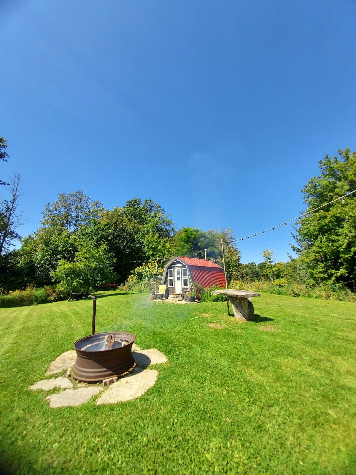 A small cabin with a red roof is set amidst a green lawn, surrounded by trees. A campfire pit, with smoke rising gently, is situated in front of the cabin. A rustic wooden table is nearby, enhancing the natural outdoor setting.