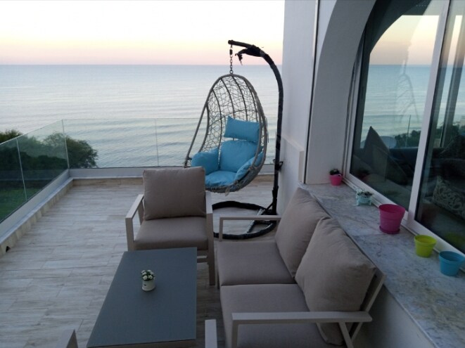 A private terrace offers stunning sea views, featuring a hanging chair with blue cushions and a light-colored outdoor seating area. A small table is present, accompanied by colorful planters. The serene atmosphere is complemented by the horizon where the sea meets the sky.