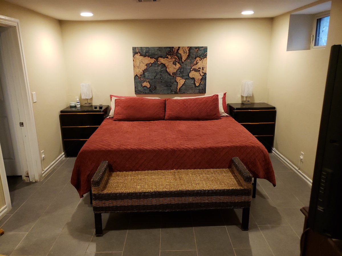 A spacious bedroom features a large bed with a textured red coverlet. Two nightstands with lamps are situated on either side. A woven bench is positioned at the foot of the bed, and a decorative world map is mounted on the wall.