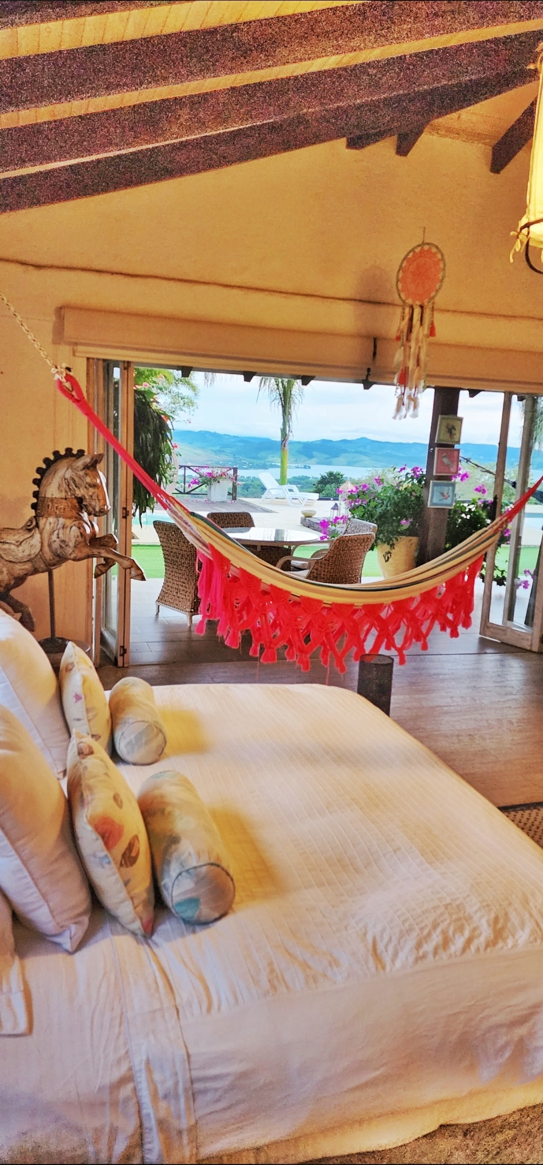 A spacious bedroom includes a large, plush bed adorned with decorative pillows. A bright hammock hangs nearby, offering a relaxed atmosphere. Sliding glass doors open to a scenic outdoor area with views of lush greenery and a body of water, enhancing the tranquil setting.