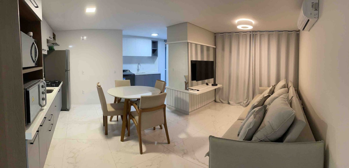 A modern living space is featured, showcasing a round dining table surrounded by four wooden chairs. A comfortable sofa is positioned adjacent to a large TV, while light-colored cabinetry and appliances are visible in the kitchenette area. Soft curtains frame the window, allowing light to filter in.