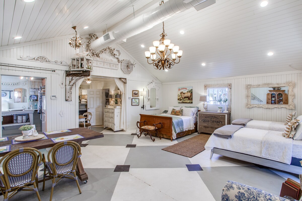 An open studio bedroom is highlighted by cathedral ceilings and chic furnishings. A queen bed and two twin beds are arranged thoughtfully, with cozy linens. A wooden dining table with chairs is positioned nearby, complemented by vintage decor on the walls.