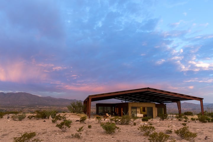 Modern Solar Home, a modest and simple structure in a desert landscape under pink and blue sky. 