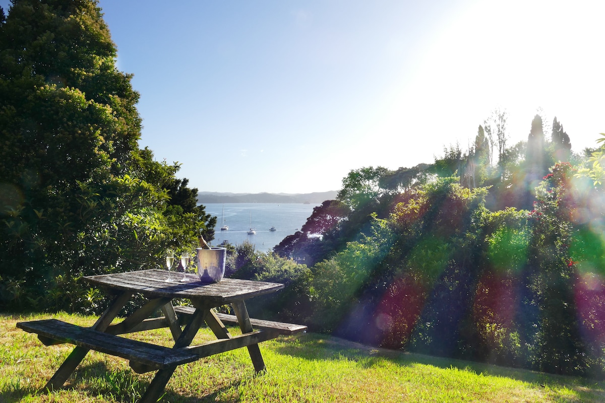 A wooden picnic table is set on a grassy area overlooking the water, with sunlight illuminating the scene. Lush greenery surrounds the table, creating a serene atmosphere with trees in the background and boats visible on the water.