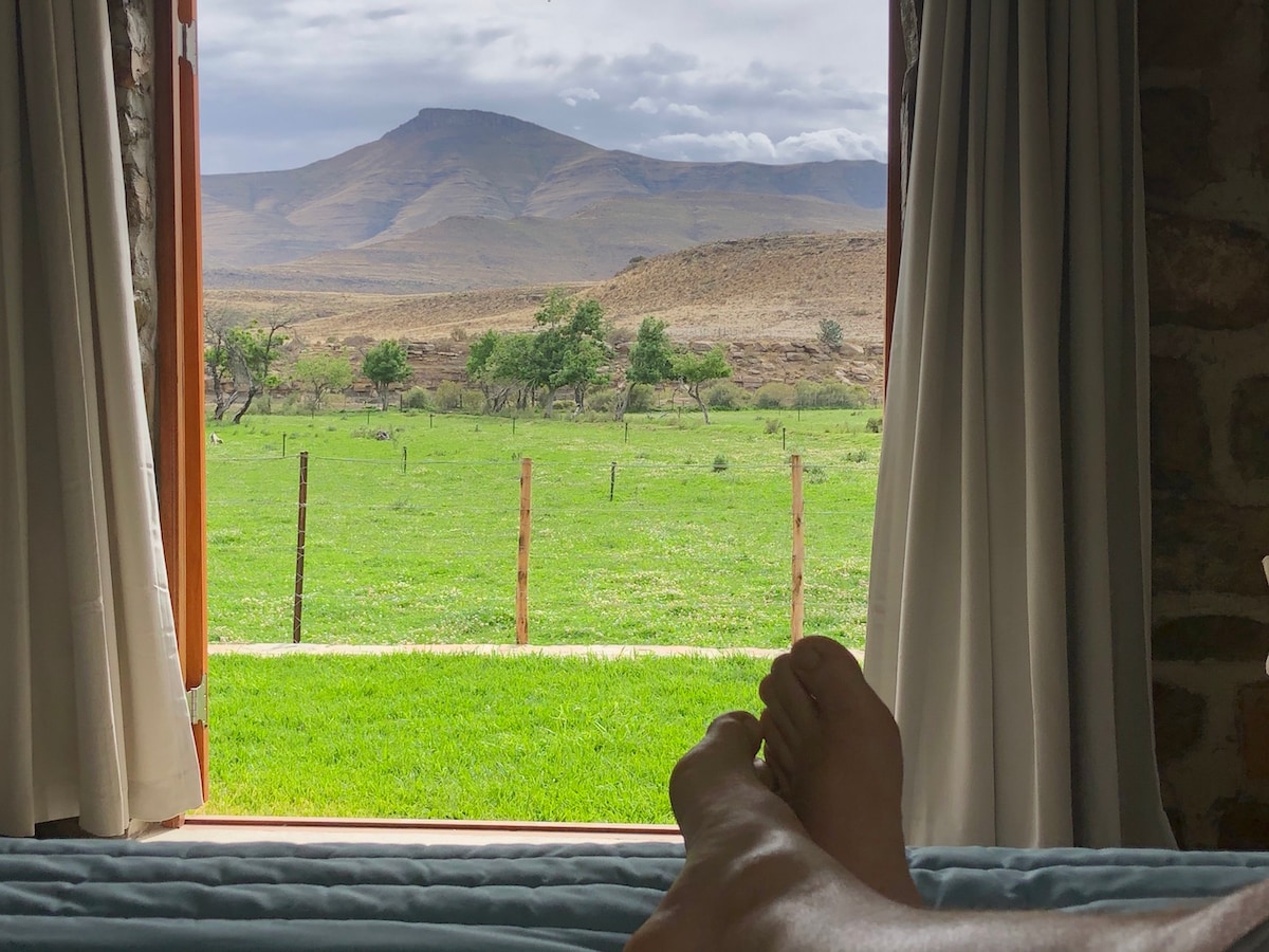 Two bare feet are visible resting on a cozy bed, with a view of lush green fields and the Sneeuberg Mountain Range framed by open curtains. The tranquil outdoor landscape showcases gentle hills under a cloudy sky.