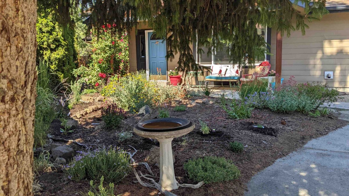 A garden area is highlighted, featuring a stone birdbath and a variety of green plants and colorful flowers. A cozy seating arrangement is visible next to the front entrance, framed by lush shrubs and trees, creating a welcoming outdoor space.