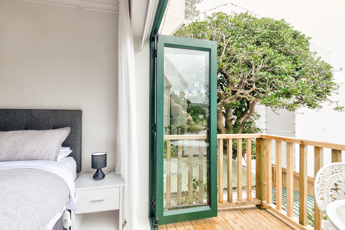 A spacious sleeping area features a comfortable bed with soft linens. A green door opens to a private deck with wooden flooring, providing a view of trees. A small lamp sits on the bedside table, contributing to the overall serene setting.