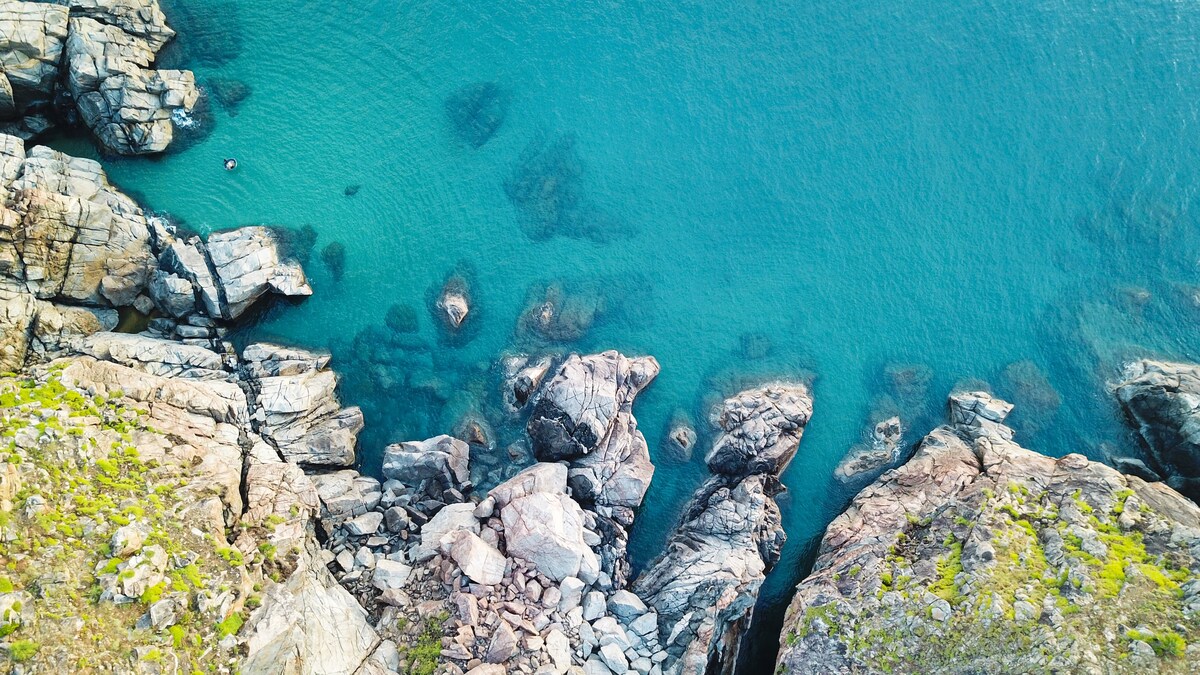 An aerial view captures the rocky coastline merging with clear blue waters. The landscape features various sizes of boulders nestled among the shoreline, while hints of greenery are visible on the rocks, creating a natural contrast against the tranquil sea.