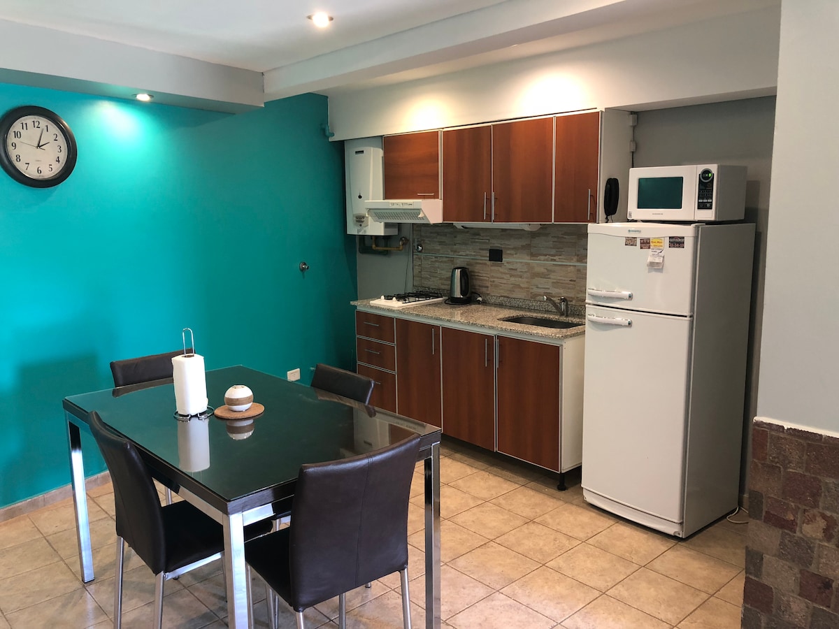 The modern kitchen area features a sleek refrigerator, microwave, and stovetop, complemented by stylish wooden cabinetry. A dining table with four chairs occupies the space, while a wall clock adds practicality. The turquoise accent wall brings a pop of color to the neutral tile flooring.