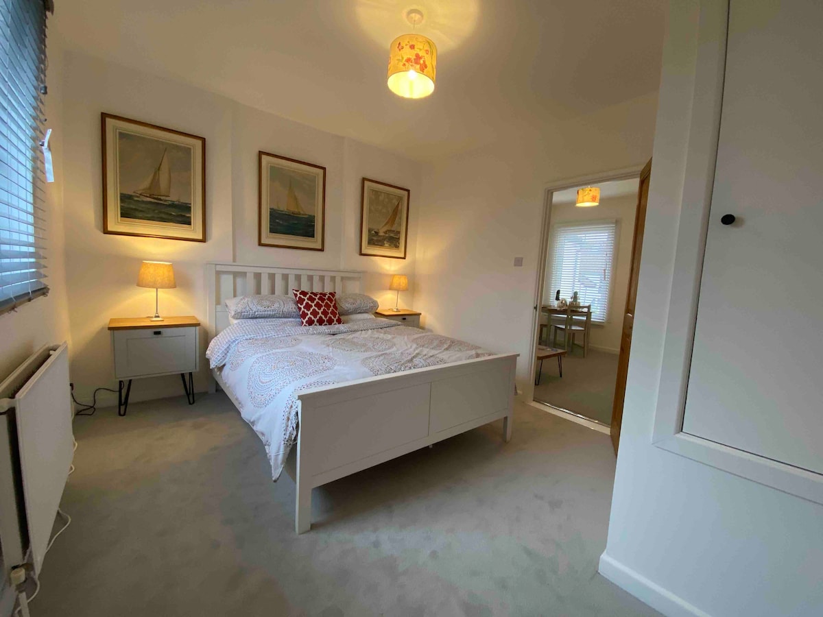 A well-lit bedroom features a spacious bed adorned with decorative pillows. Two bedside lamps provide soft lighting, while framed nautical artworks decorate the walls. A door leads to the dining area, and a built-in wardrobe is visible on the right.