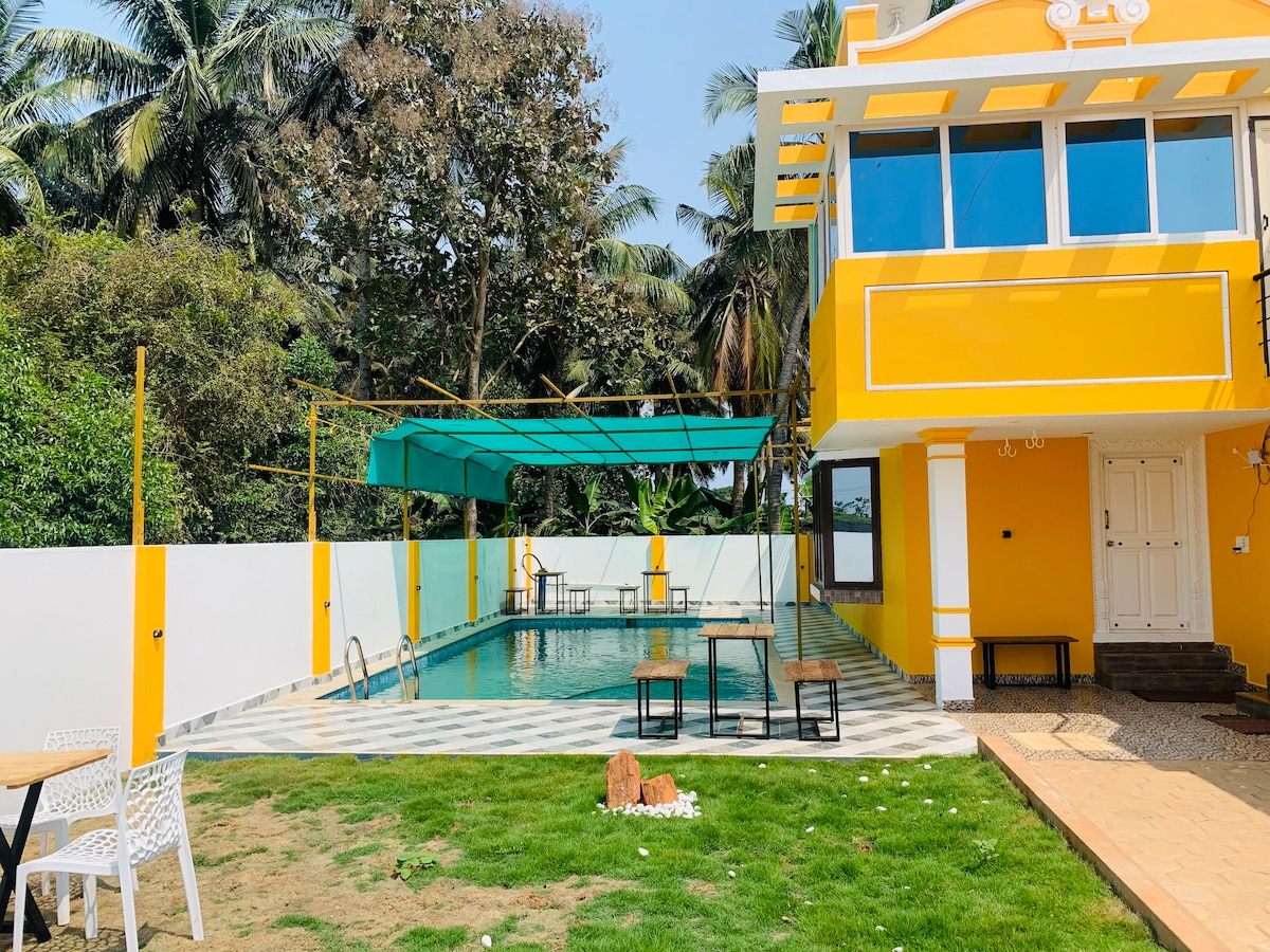 A vibrant villa exterior features a spacious swimming pool surrounded by a neatly maintained lawn. Two tables are placed on the grassy area, complemented by a shaded seating area. Lush greenery and palm trees provide a serene backdrop against the cheerful yellow structure.