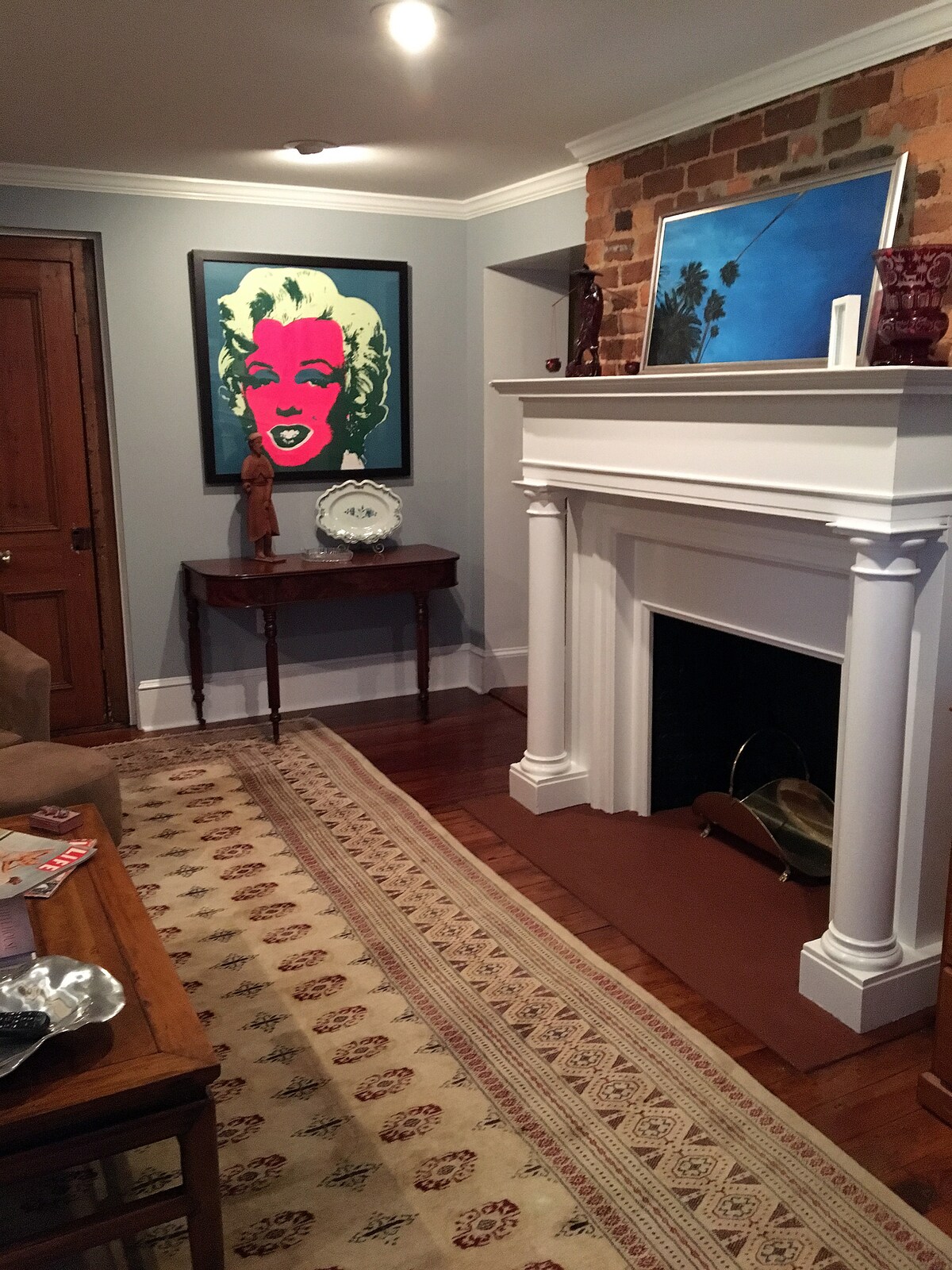 A cozy living area showcases a vibrant piece of artwork featuring a famous figure, positioned above a classic white mantle. An antique table stands adjacent to the fireplace, with light-colored wood floors and a patterned rug enhancing the space. Subtle wall textures create a warm backdrop.