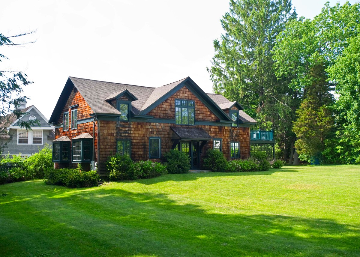 A charming exterior of the carriage house features rustic wooden shingles and large windows, surrounded by well-maintained landscaping. Lush green grass extends towards the home, complemented by mature trees in the background, creating a serene outdoor setting.