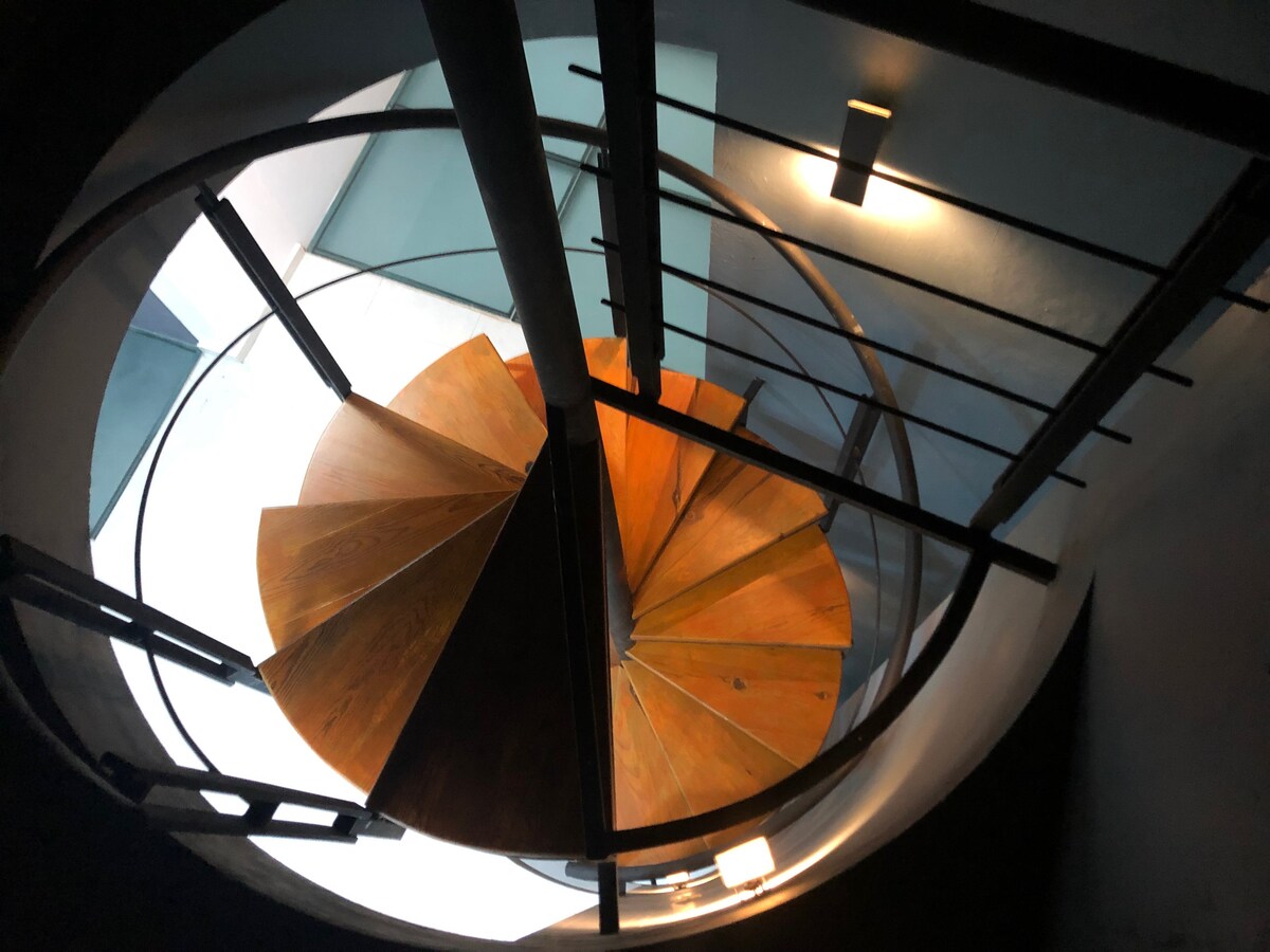 An elegantly designed spiral staircase is showcased, constructed of wood and surrounded by metal railings. The staircase leads to an open area illuminated by natural light, creating a sense of connection between the levels of the home.