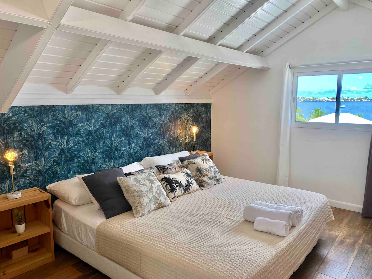 A bright bedroom features a spacious bed with an array of decorative pillows. The wall behind is adorned with tropical-themed wallpaper. Natural light streams in through the window, which offers a view of the exterior landscape, accentuating the airy atmosphere.