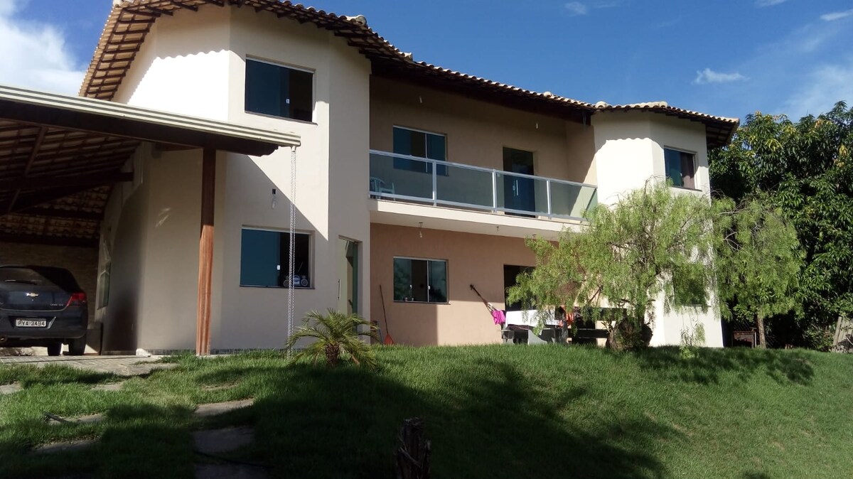 The two-story building features a spacious balcony with glass railings, providing views of the surrounding greenery. Large windows allow natural light to fill the interiors. A carport is visible in the foreground, and well-maintained grass leads up to the entrance.
