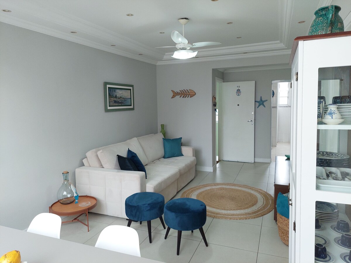 A bright living area features a comfortable sofa bed paired with two accent chairs. A ceiling fan contributes to airflow in the space. An artwork adorns the wall, while a light-colored dining table awaits nearby, enhancing the overall openness of the room.