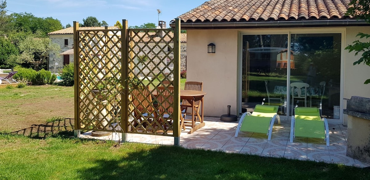 An outdoor patio area features a wooden trellis, creating a sense of privacy. Light green lounge chairs are arranged beside a small table, surrounded by lush grass. The patio opens to a view of the serene landscape, with a glimpse of a pool in the background.