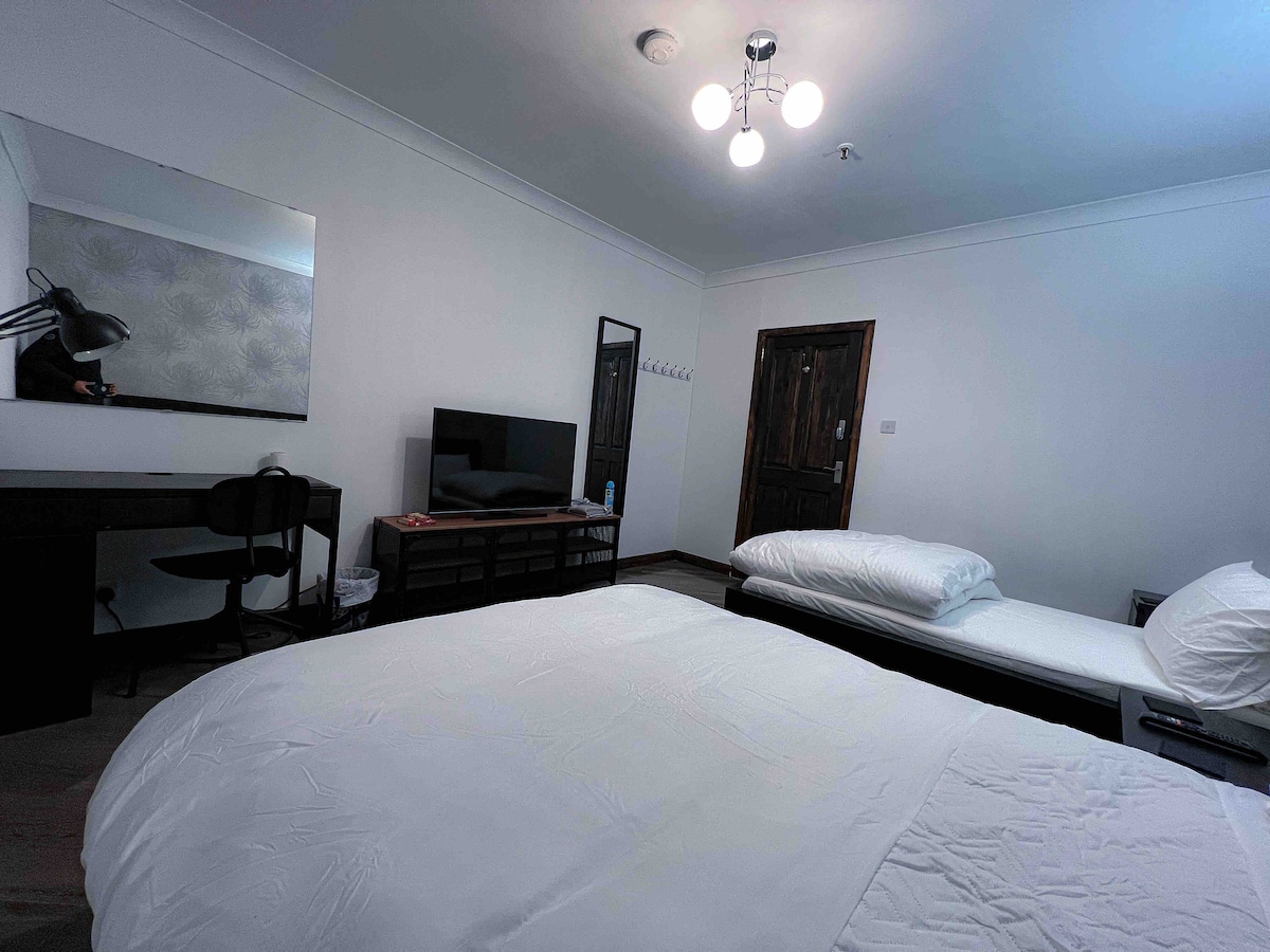 A spacious bedroom features a comfortable double bed with crisp white linens alongside a single bed. A desk with a lamp and a television are visible, complemented by a mirror and a wardrobe. Soft lighting is provided by a modern ceiling fixture.