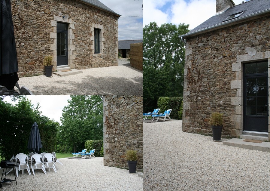 The exterior of the gîte features rustic stone walls and a gravel courtyard. Multiple seating options, including white chairs and loungers, are arranged around the space. A single umbrella provides shade. Lush greenery surrounds the area, enhancing the outdoor ambiance.