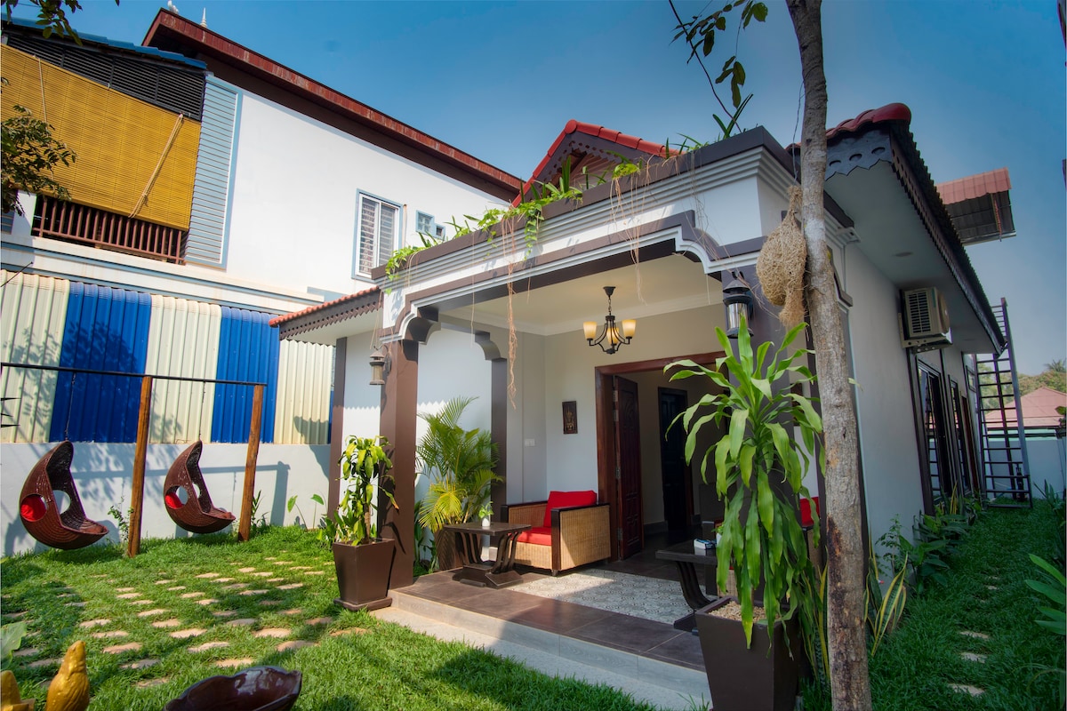 The exterior showcases a harmonious blend of Khmer and modern architecture. A welcoming porch features comfortable seating, surrounded by greenery and a manicured lawn. Two hanging chairs invite relaxation, while a classic chandelier adds a touch of elegance to the inviting entrance.