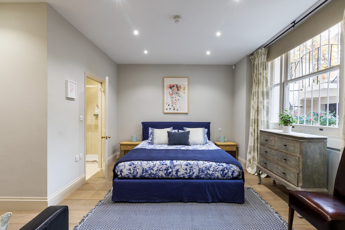 The spacious bedroom features a large king-size bed with a blue floral duvet. Natural light fills the room through two large windows, complemented by soft curtains. A small art piece hangs on the wall, and a decorative chest of drawers is placed nearby.