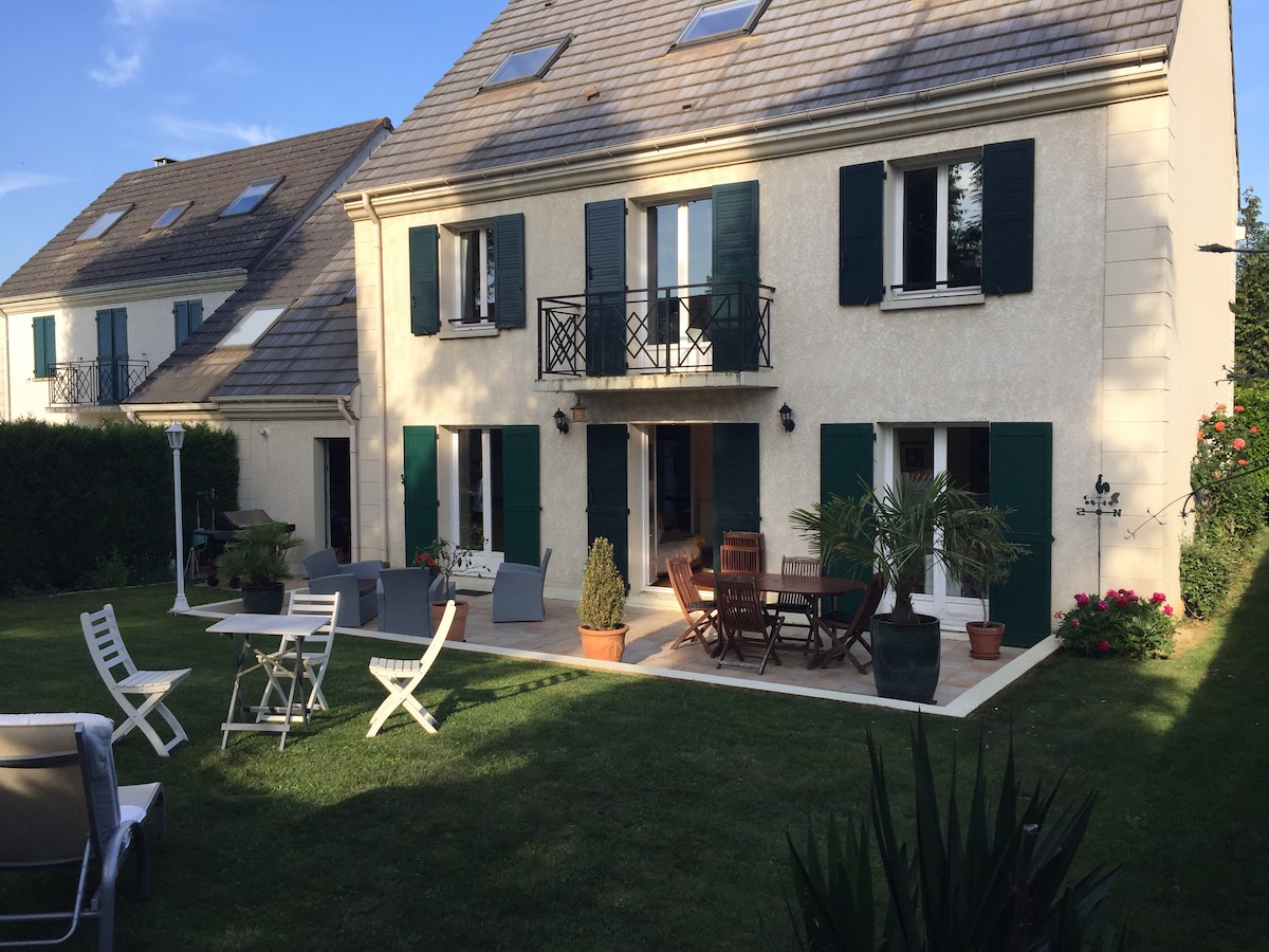 A spacious house is shown, featuring green shutters and multiple balconies. A well-maintained garden area is visible, with patio furniture arranged for outdoor dining and relaxation. Sun loungers and potted plants are scattered across the lawn, complementing the serene setting.